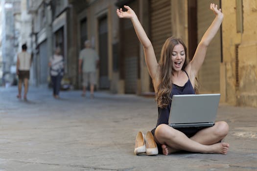 Gig economy freelancer A cheerful woman sitting outdoors, celebrating success with arms raised, while using a laptop.