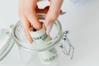 How to build a budget that works. A close-up of a hand placing rolled dollars into a glass jar, symbolizing savings.