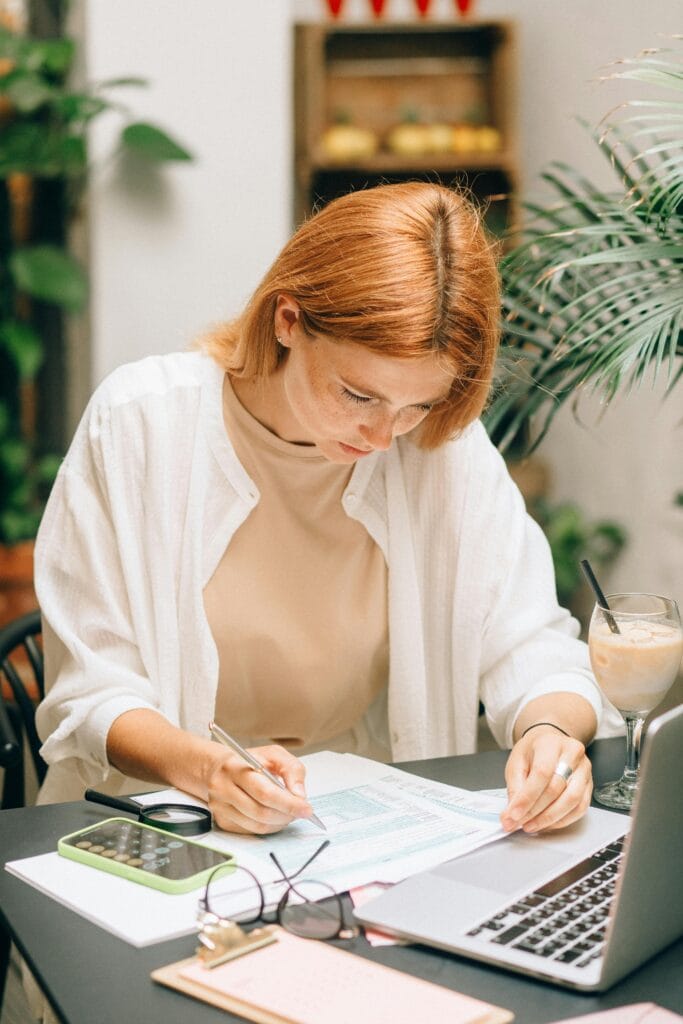 How to build a budget that works, Woman analyzing financial documents using laptop and calculator indoors.