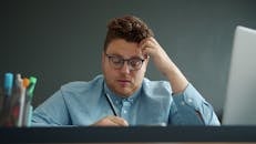 Man in glasses focusing on writing at his desk with a pensive look.
